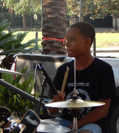 Young boy playing drum set outdoors during the daytime beside greenery and parked cars.