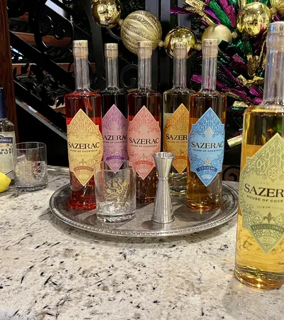 Six colorful Sazerac cocktail liquor bottles arranged on a silver tray with glasses, jigger, syrup, and a lemon on marble countertop.