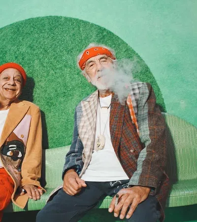Two stylish seniors in vibrant outfits relaxing on a green bench with smoke in the air.