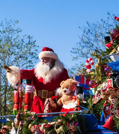 Santa Claus waves from a festive blue parade float decorated with toys, Christmas garlands, and candy canes under clear skies.