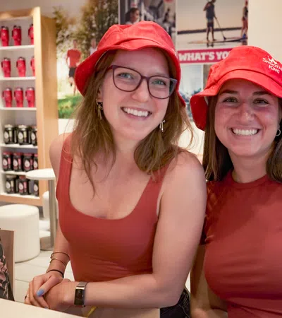 Two smiling women wearing red shirts and red hats sitting indoors with shelves and posters in the background