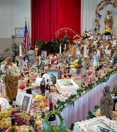 Indoor religious altar with statues, candles, flowers, food offerings, and people attending a celebration.