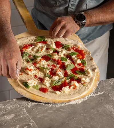 Person preparing homemade pizza with tomato sauce, cheese, and basil on a wooden paddle.