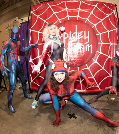 Four cosplayers dressed as Spider-Man characters pose dynamically in front of a red spider web backdrop at a convention.