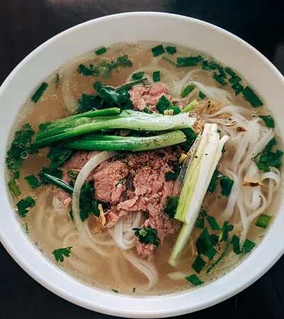 Bowl of Vietnamese pho with beef, rice noodles, green onions, and herbs in clear broth on dark surface
