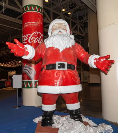 Life-size Santa Claus statue with open arms near Coca-Cola branded pillar at an indoor event.