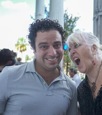 Smiling man and woman with open mouth caught mid-shout outdoors by stone columns and greenery in background