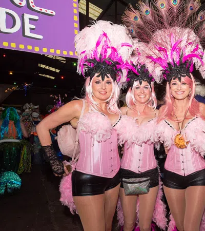 Four women in pink feathered showgirl costumes with black accents posing indoors at a festive event.