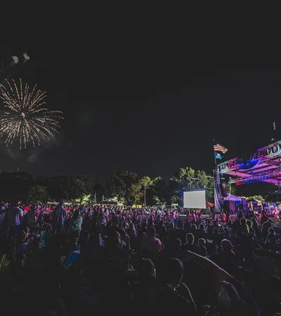 Crowd at outdoor night concert with fireworks exploding in the sky above the stage and audience