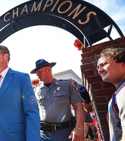 Man in blue suit walking under Walk of Champions arch with crowd and police officer nearby