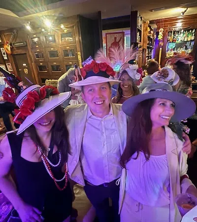Group of smiling people wearing colorful hats enjoying a lively indoor party with drinks and wooden decor.
