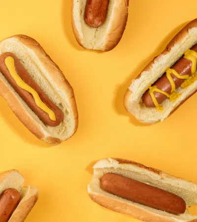 Several hot dogs in buns on a yellow background, some topped with yellow mustard in various patterns.