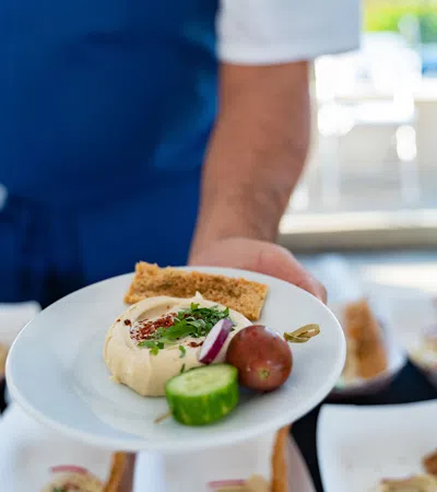 Chef holding plate with hummus, pita bread, cucumber slice, grape tomato, and garnish in a kitchen setting