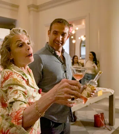 Older woman pouring wine while smiling man watches in elegant room with guests and decorations