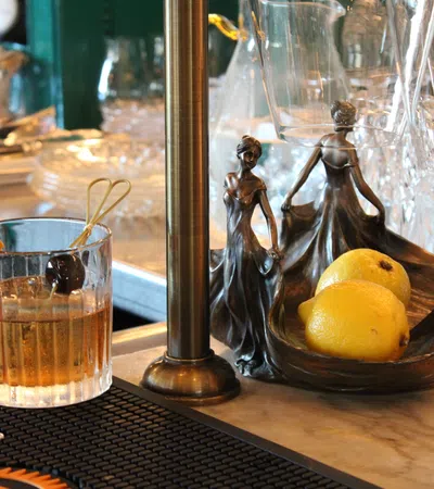 Whiskey cocktail with orange peel and cherry next to an art deco lemon holder shaped as dancing ladies on bar counter.