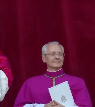 Pope in ornate vestments waving with two clergymen in ceremonial robes against red curtain background.