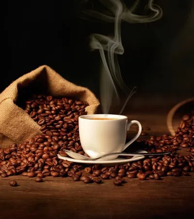 Steaming cup of coffee surrounded by spilled coffee beans from burlap sacks on a wooden surface.