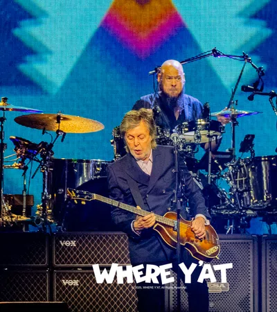 Paul McCartney playing bass guitar with drummer behind him on stage during a concert with a colorful background.