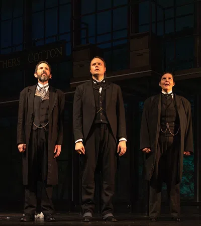 Three men in vintage suits stand on stage in front of an industrial backdrop under warm lighting during a theater performance.