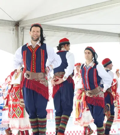 Group of dancers performing traditional folk dance in colorful Eastern European costumes under a white tent.
