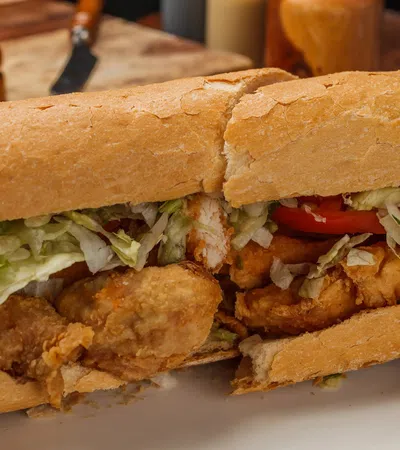 Crispy fried chicken po'boy sandwich with lettuce and tomato on a crusty French bread roll on a white plate.