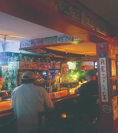 Interior of a cozy bar with patrons seated at the wooden counter, vintage signs, and warm lighting.