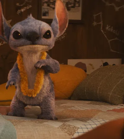 Blue alien creature wearing a yellow lei standing on a bed in a cozy room with photos and a stuffed dog toy.