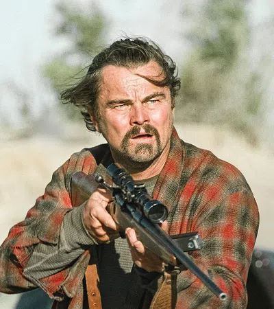 Man aiming a scoped hunting rifle outdoors wearing a red plaid shirt with intense focused expression.