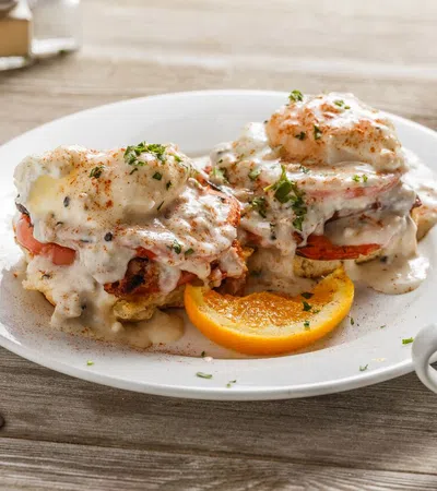 Plate of eggs Benedict with creamy sauce and herbs, garnished with an orange slice, next to a cup of black coffee.
