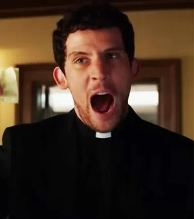Man in black clerical collar raising finger and shouting while older man in tan suit watches in room.