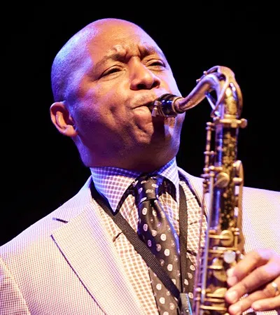 Jazz musician passionately playing saxophone on stage in a light-colored suit and patterned tie.