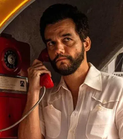 Man with beard using a red vintage phone booth with political posters on the wall behind him.