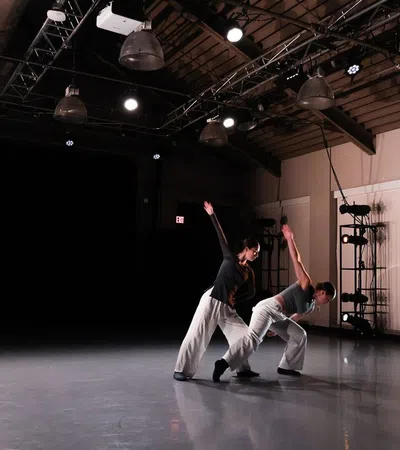 Two contemporary dancers performing in a dimly lit studio with spotlight and industrial ceiling beams.