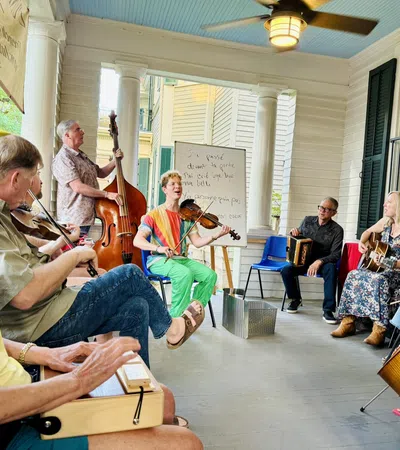 Group of people playing various musical instruments together on a covered porch during daylight.