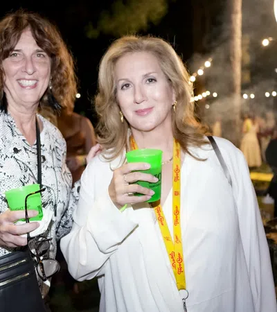 Two women smiling and holding green cups at an outdoor nighttime social event with string lights in background