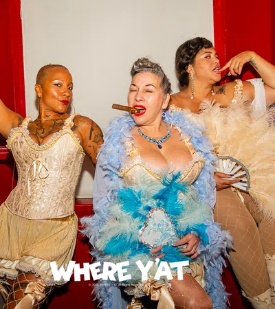 Three women in vintage burlesque costumes pose with feathers and a cigar against a red and white backdrop.