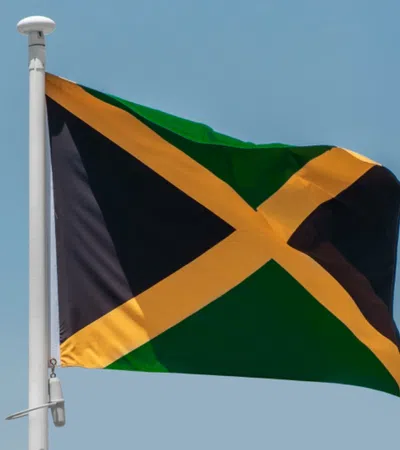 Jamaican flag waving on a white flagpole against a clear blue sky background.