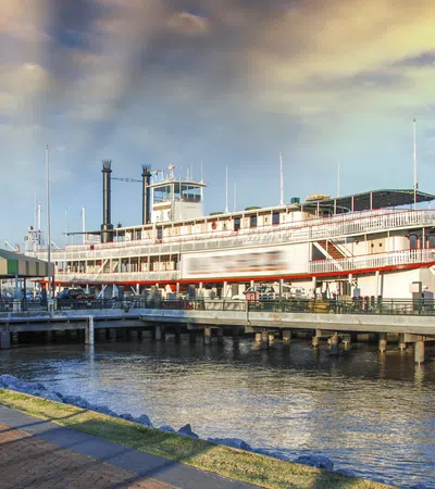 Historic riverboat docked at pier with red paddlewheel under a partly cloudy sky at sunset.