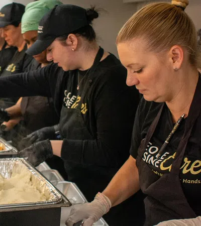 Volunteers wearing black gloves and aprons serve food from large trays in a community meal line.