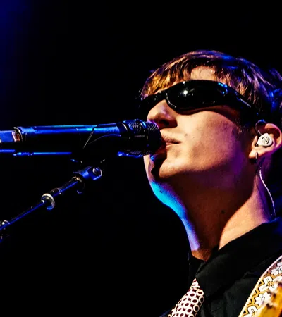 Male musician wearing sunglasses singing into microphone on stage with guitar under blue lighting