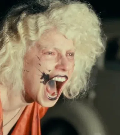 Person with wild blonde hair and black paint splatter on face yelling with mouth wide open in intense expression