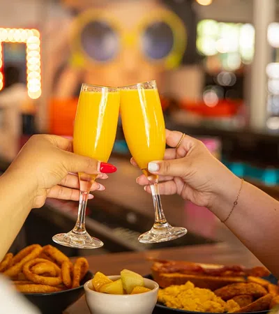 Two people clinking glasses of orange mimosa cocktails over a table with snacks in a cozy bar setting