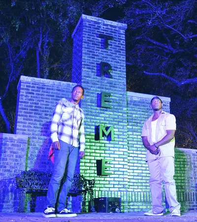 two men standing in front of a building with a fence and trees