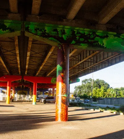 Colorful mural of trees painted on the columns supporting a highway overpass in a sunny urban area.