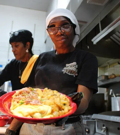Smiling chef in kitchen holding a red plate with breakfast food including eggs, potatoes, and bacon.