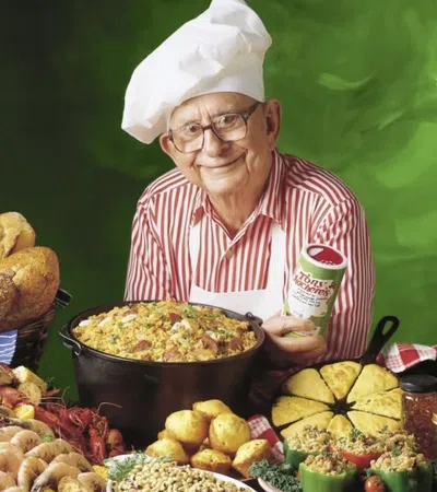 Elderly chef in white hat and red striped shirt presenting a large dish surrounded by bread, seafood, and vegetables.