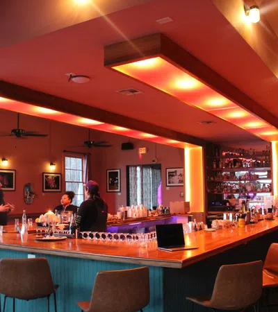 Modern bar interior with warm lighting, patrons chatting at L-shaped wooden counter, and high chairs surrounding it.