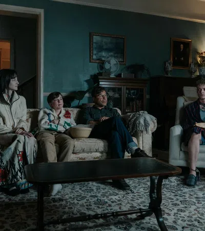 Four people sitting in a dimly lit living room, one chained to a chair holding a bowl.