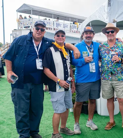 Group of eight men wearing colorful shirts and hats posing on green turf at an outdoor event under a white tent.