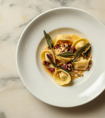 White plate with gourmet tortellini pasta garnished with sage leaves, nuts, and sauce on a marble surface.